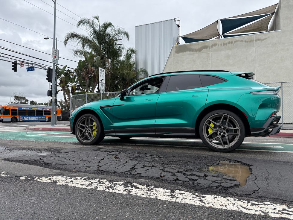 A turquoise luxury SUV parked on a city street featuring visible potholes and urban signage.