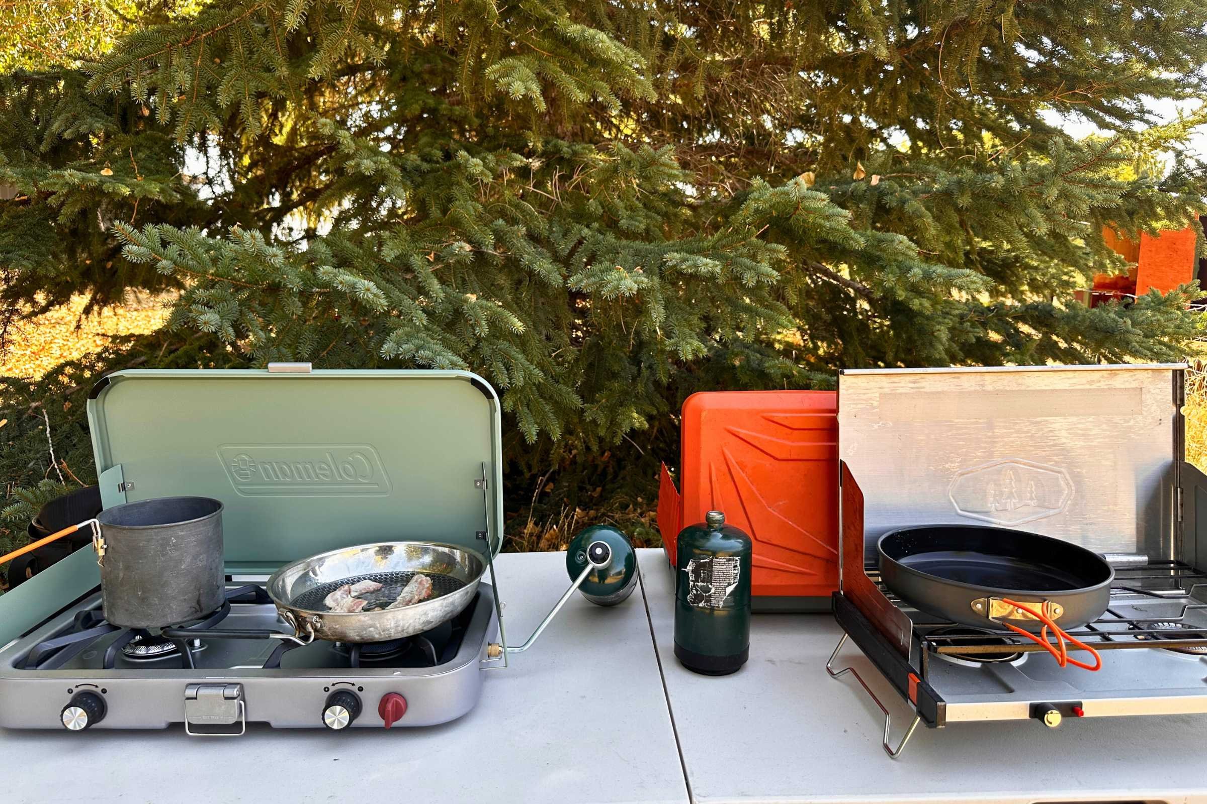 Two camping stoves neatly set up on a wooden table in a forest clearing.