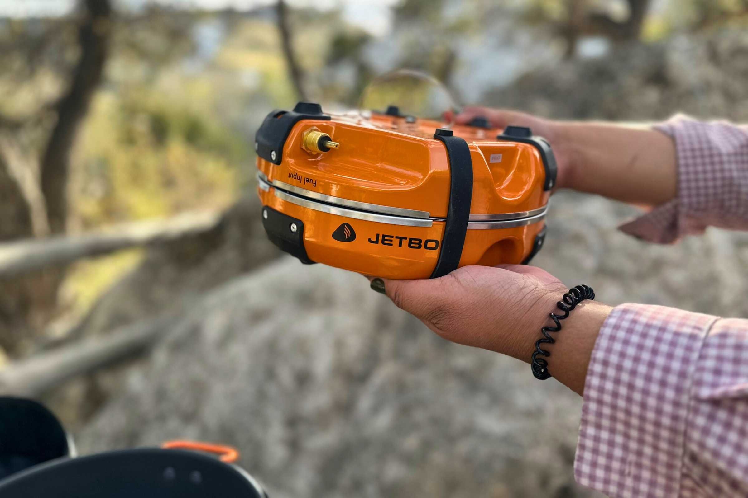 Hands holding a folded orange Jetboil Genesis stove to demonstrate its compact size.