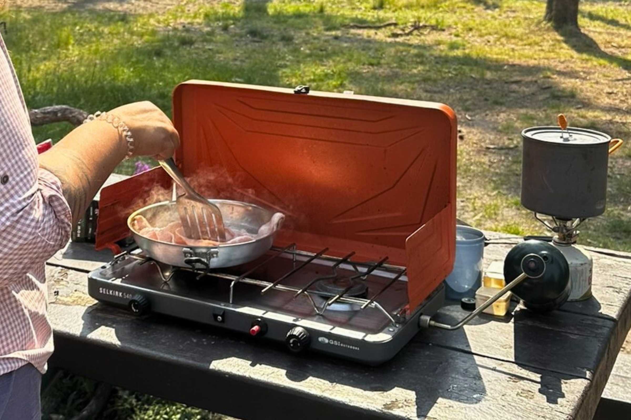 An outdoor chef preparing breakfast on a GSI stove at a wooden picnic table.