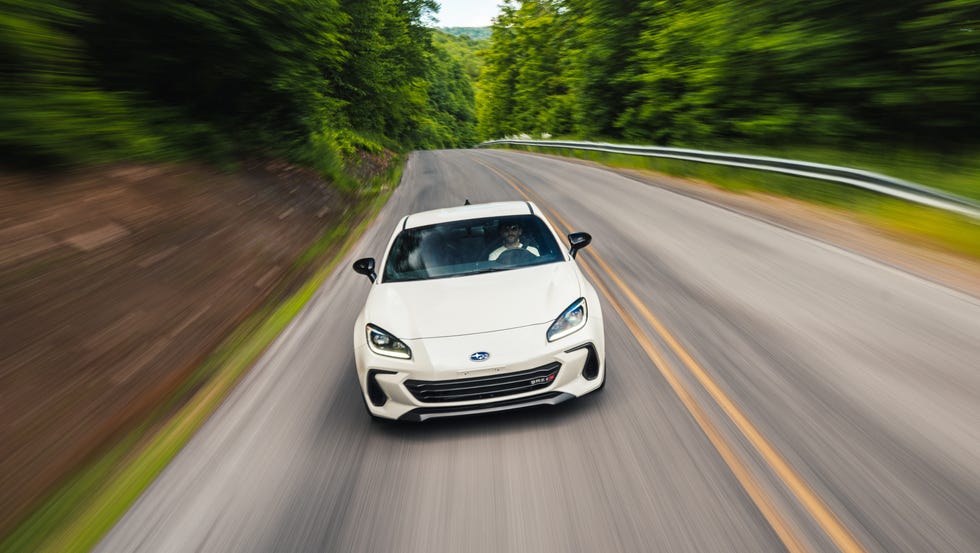 A white Subaru BRZ tS driving through a sharp, forested corner on a winding road.