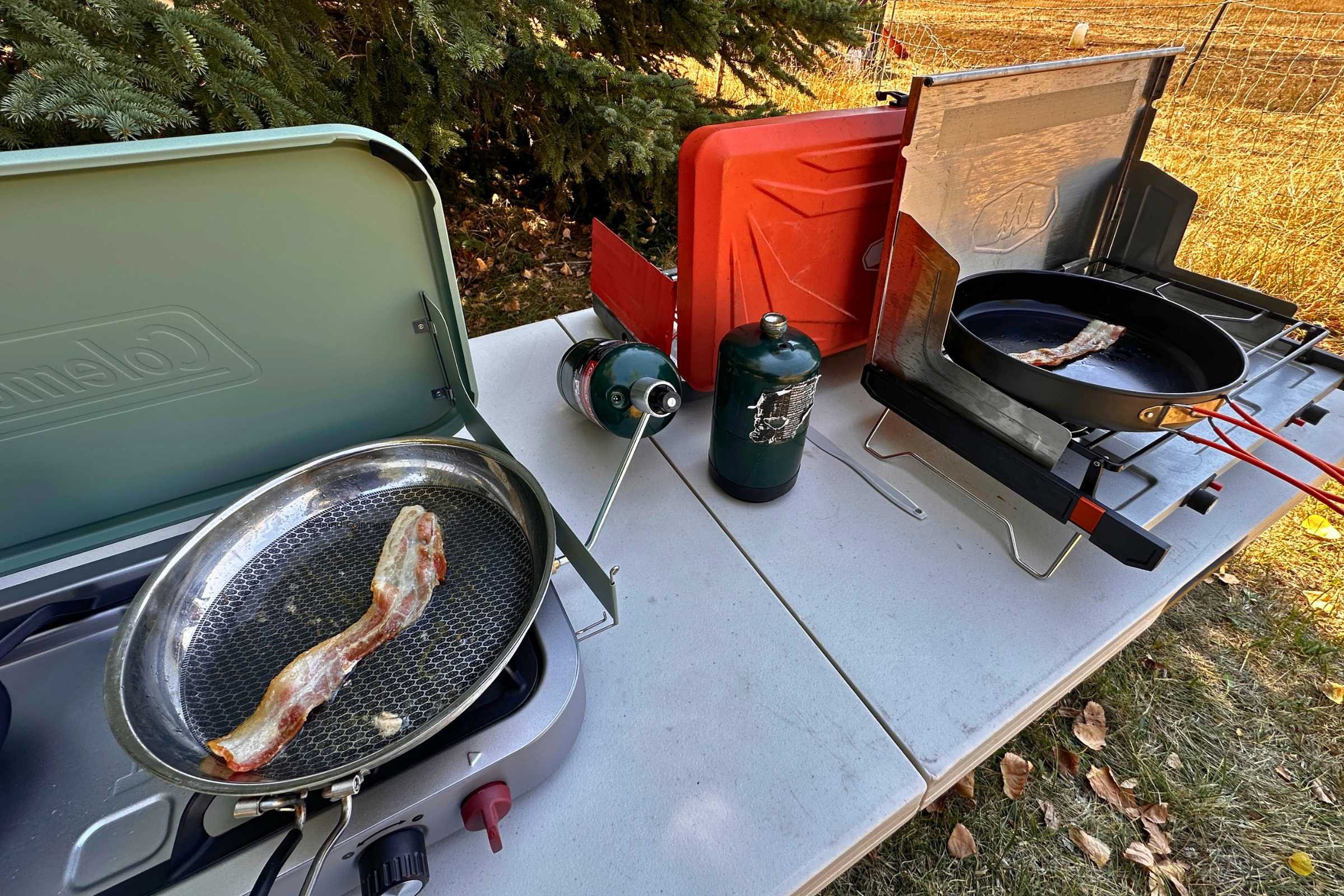 Multiple camping stoves being tested side-by-side with bacon cooking.