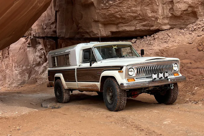 A 1975 Jeep J10 with a vintage Caravan Camper shell parked in a desert landscape.