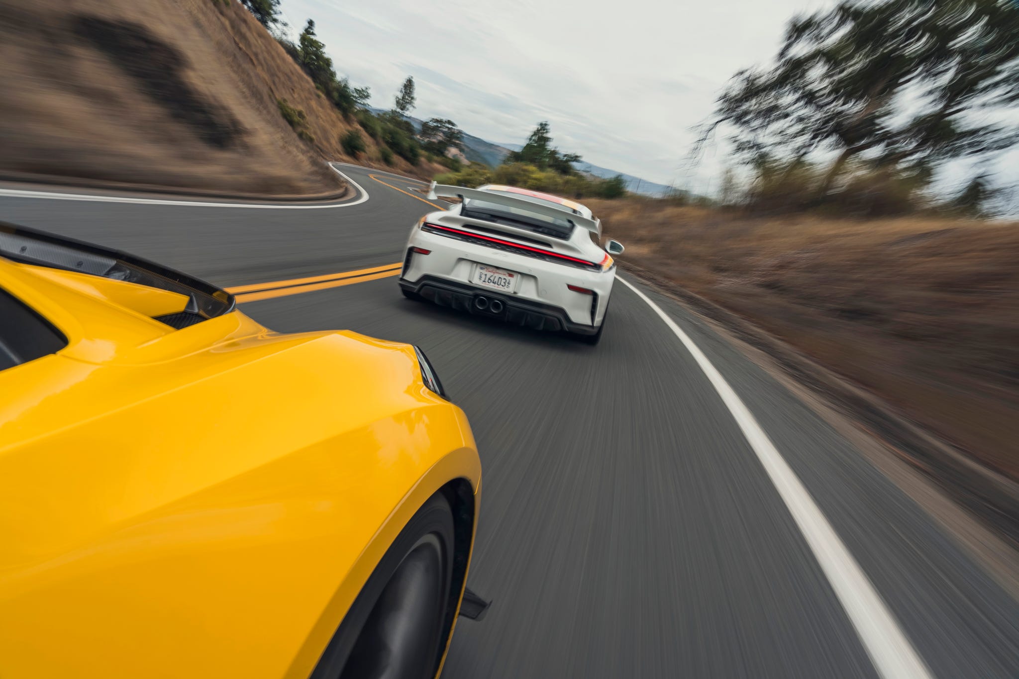 A yellow Chevrolet Corvette ZR1 followed by a blue Porsche 911 GT3 on a narrow, winding mountain road.